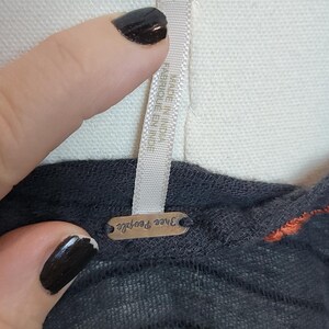Puede incluir: Primer plano de una camiseta gris oscuro con rayas horizontales y un acento naranja. La etiqueta dice "Free People". La camiseta est&aacute; sobre un maniqu&iacute; blanco.
