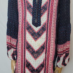 May include: A long-sleeved tunic dress with a V-neckline. The dress features a navy blue background with a pattern of small white dots. The front panel has a chevron design in white, pink, and navy blue.