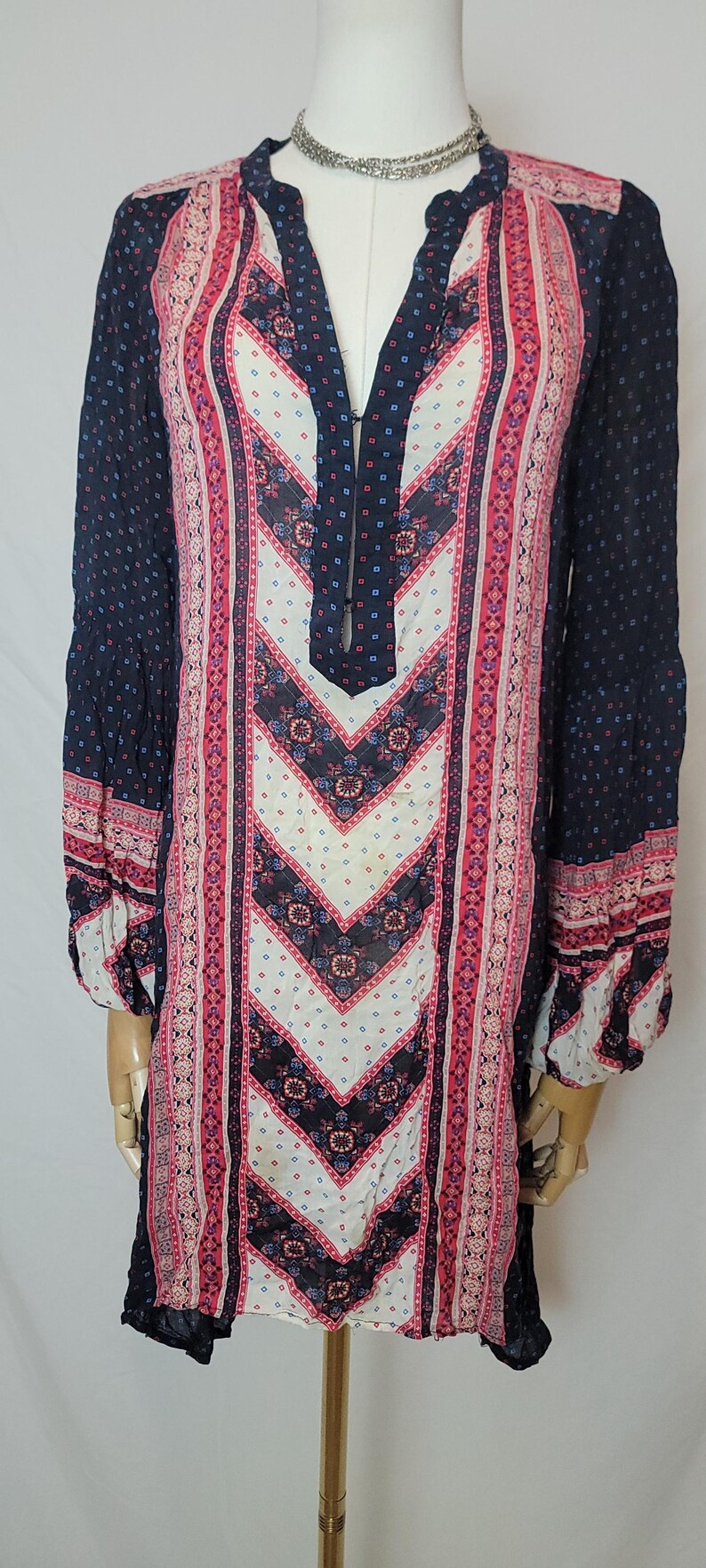 May include: A long-sleeved tunic dress with a V-neckline. The dress features a navy blue background with a pattern of small white dots. The front panel has a chevron design in white, pink, and navy blue.