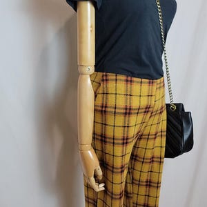K&ouml;nnte beinhalten: Eine Schaufensterpuppe zeigt ein schwarzes T-Shirt und eine gelbe karierte Hose mit einer schwarzen Handtasche. Die Hose hat eine senfgelbe Basis mit einem rot-schwarzen Karomuster. Die Handtasche hat einen Kettenriemen.