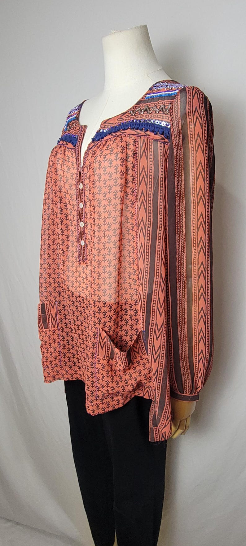 Puede incluir: Blusa color coral con estampado floral y rayas verticales marrones. La blusa tiene cuello en V, detalles de botones y bolsillos. El escote y las mangas est&aacute;n adornados con adornos azules y coloridos.
