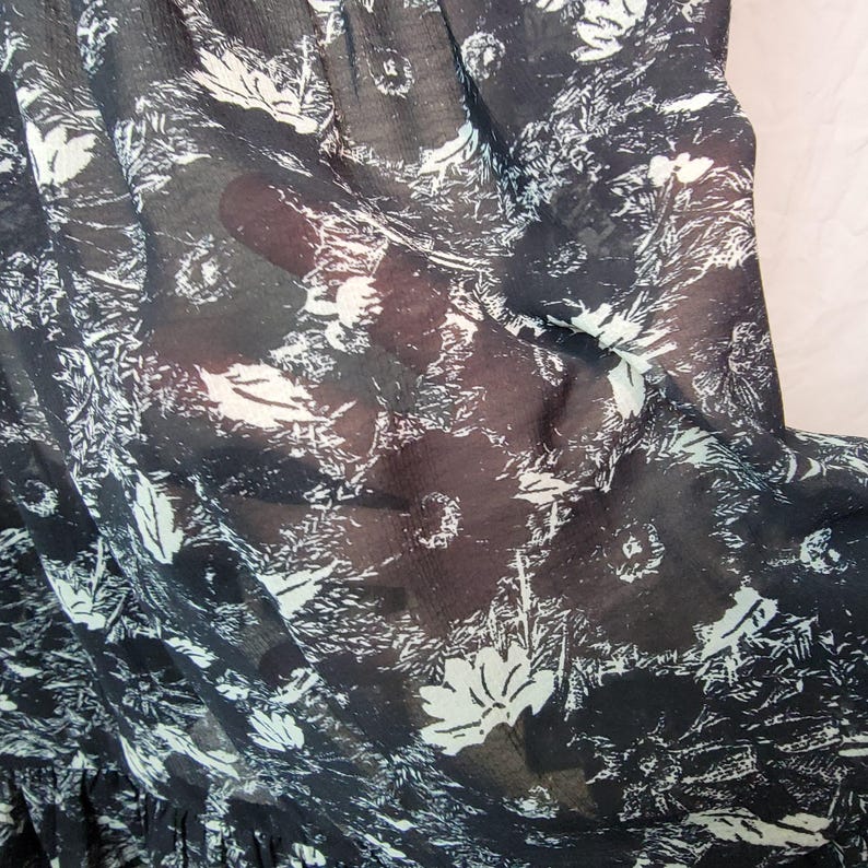 Puede incluir: Primer plano de una tela negra transparente con un estampado floral blanco. La tela es ligera y parece ser un material de vestido o blusa. El dise&ntilde;o incluye flores y follaje estilizados, creando una est&eacute;tica delicada y aireada.