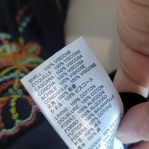 May include: A white clothing label with text in several languages, detailing the fabric composition. The label indicates the garment's shell is 100% viscose, with lining and embroidery information also provided. The tag is held by a hand.