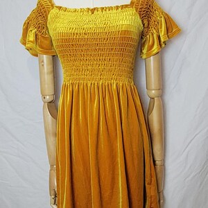 May include: Mustard yellow velvet dress with a square neckline and ruffled sleeves. The bodice is smocked, and the skirt is a-line. A gold-toned necklace is around the mannequin's neck. The dress is displayed on a wooden mannequin.