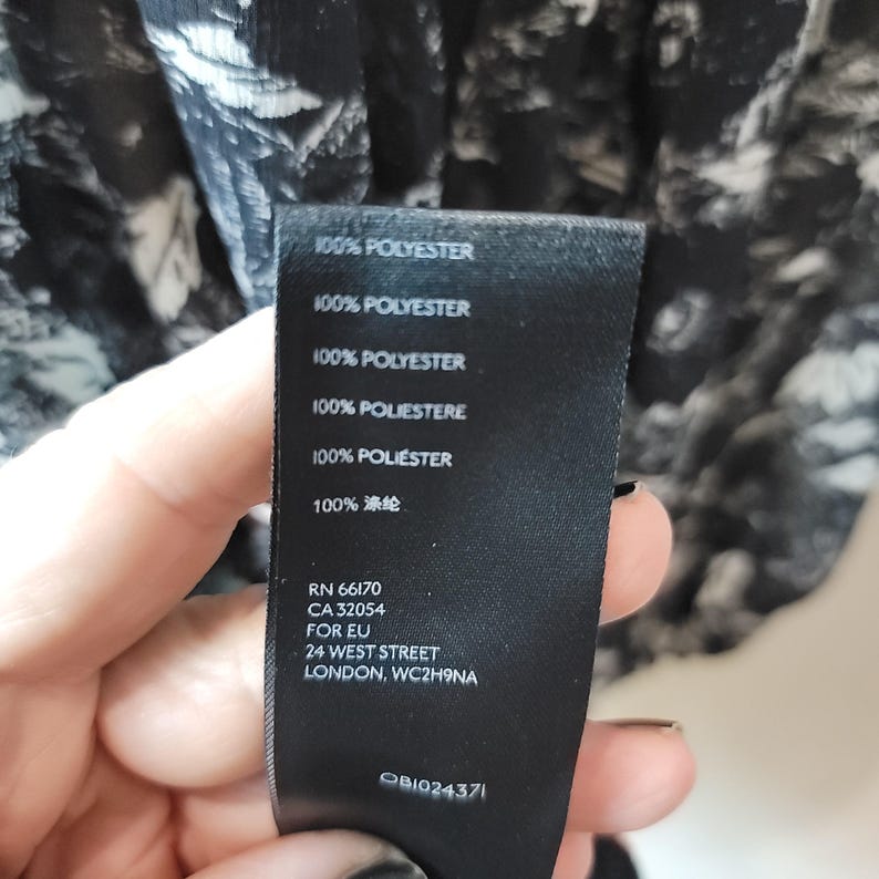 Puede incluir: Una etiqueta negra con texto blanco que indica la composici&oacute;n de la tela como 100% poli&eacute;ster. La etiqueta tambi&eacute;n incluye un n&uacute;mero de producto y la direcci&oacute;n 24 West Street, Londres, WC2H9NA.