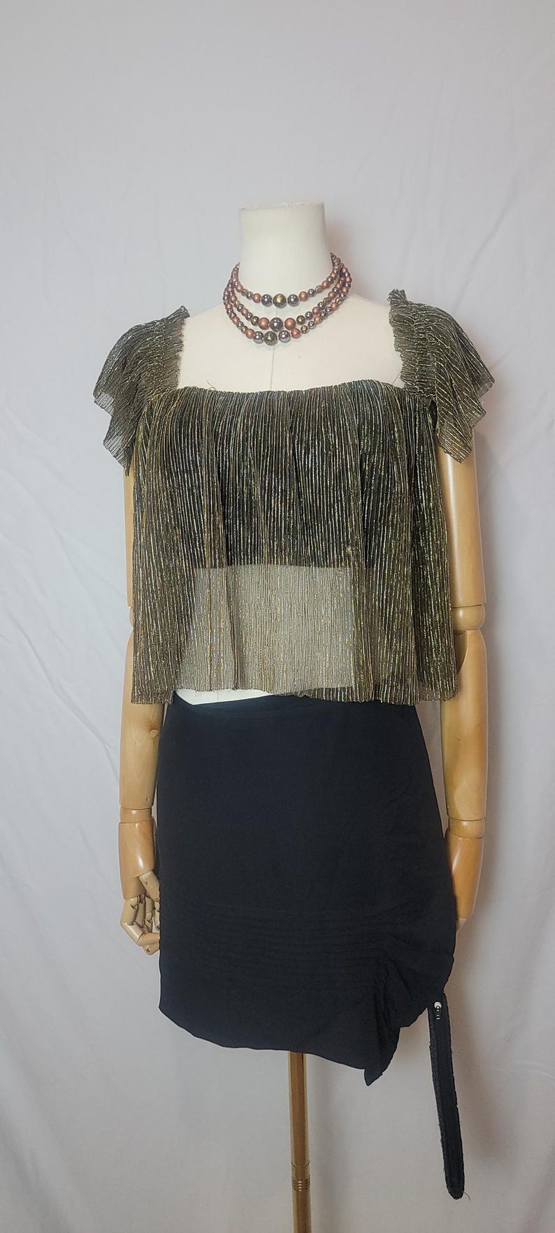 May include: A gold and black metallic crop top with a square neckline and short sleeves is displayed on a mannequin. The top is paired with a black mini skirt. A multi-strand beaded necklace is also included.