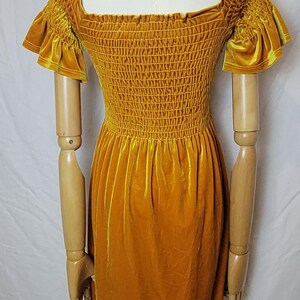 May include: Mustard yellow velvet dress with a square neckline and short, ruffled sleeves. The bodice is smocked, and the skirt is a simple A-line. The dress is displayed on a mannequin with wooden arms.