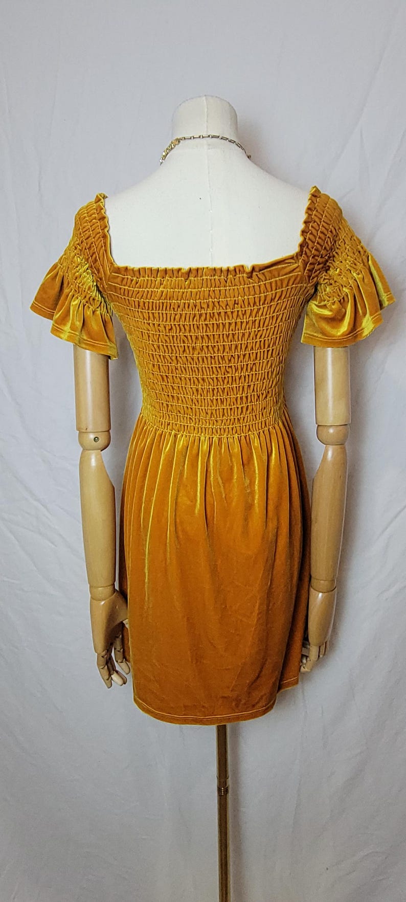 May include: Mustard yellow velvet dress with a square neckline and short, ruffled sleeves. The bodice is smocked, and the skirt is a simple A-line. The dress is displayed on a mannequin with wooden arms.