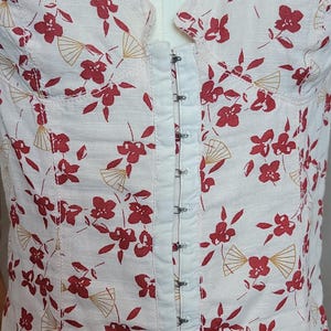 Puede incluir: Top bustier blanco con un estampado floral y abanicos rojos. El top presenta un cierre de corchetes en la parte delantera y se muestra en un maniqu&iacute;. La tela tiene un aspecto texturizado.