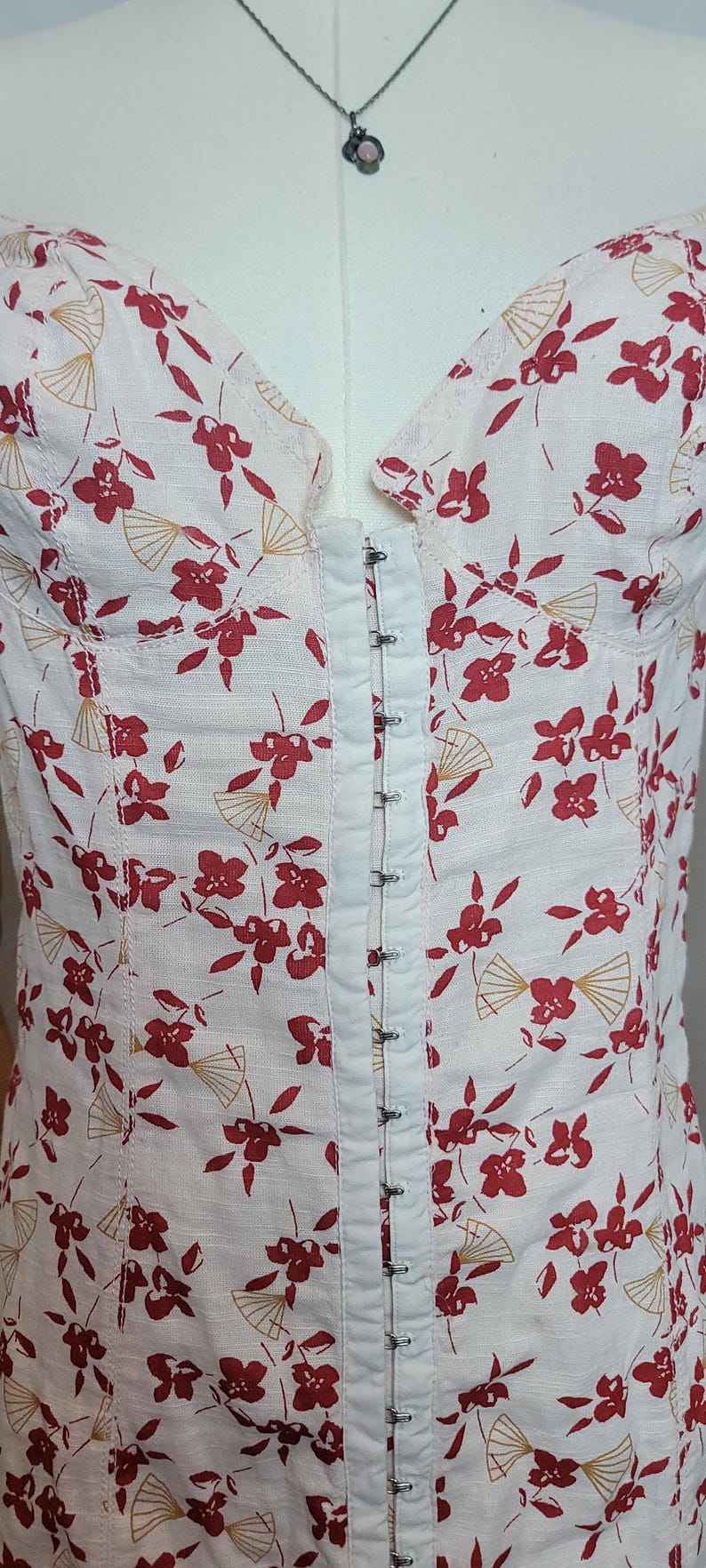 Puede incluir: Top bustier blanco con un estampado floral y abanicos rojos. El top presenta un cierre de corchetes en la parte delantera y se muestra en un maniqu&iacute;. La tela tiene un aspecto texturizado.