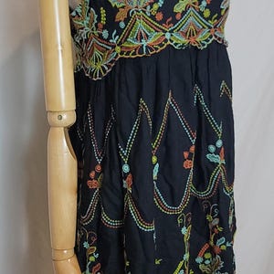 May include: Black embroidered tank top with colourful floral and geometric patterns. The top has a V-neck and thin straps. The embroidery includes orange, green, and yellow details. The garment is displayed on a mannequin.