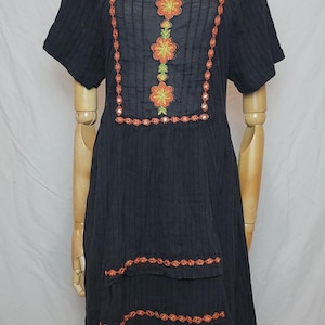 Puede incluir: Vestido negro de manga corta con bordado floral. El vestido presenta bordados florales en naranja y verde alrededor del escote y en la parte delantera. Bordados adicionales acent&uacute;an el dobladillo escalonado. El vestido se muestra en un maniqu&iacute;.