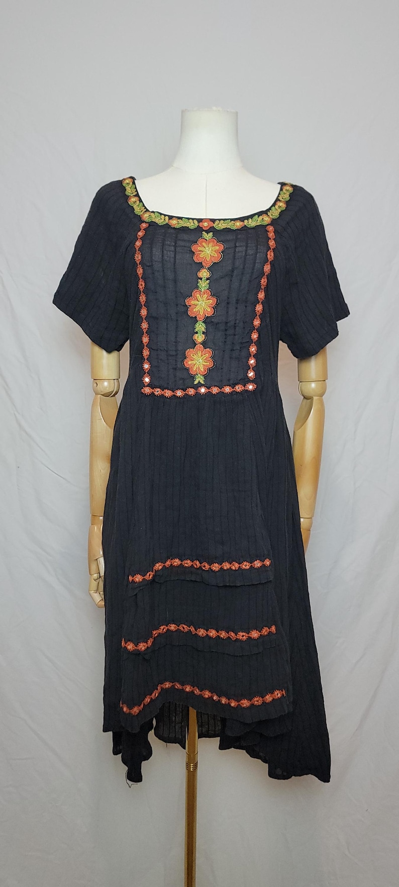 Puede incluir: Vestido negro de manga corta con bordado floral. El vestido presenta bordados florales en naranja y verde alrededor del escote y en la parte delantera. Bordados adicionales acent&uacute;an el dobladillo escalonado. El vestido se muestra en un maniqu&iacute;.