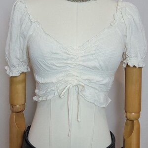 Op de afbeelding: Witte, gerimpelde crop top met korte mouwen en een trekkoord. Gedragen met een zwarte jeans en een zwarte riem met een gouden gesp. Een zilveren ketting is ook zichtbaar. De outfit wordt op een paspop getoond.
