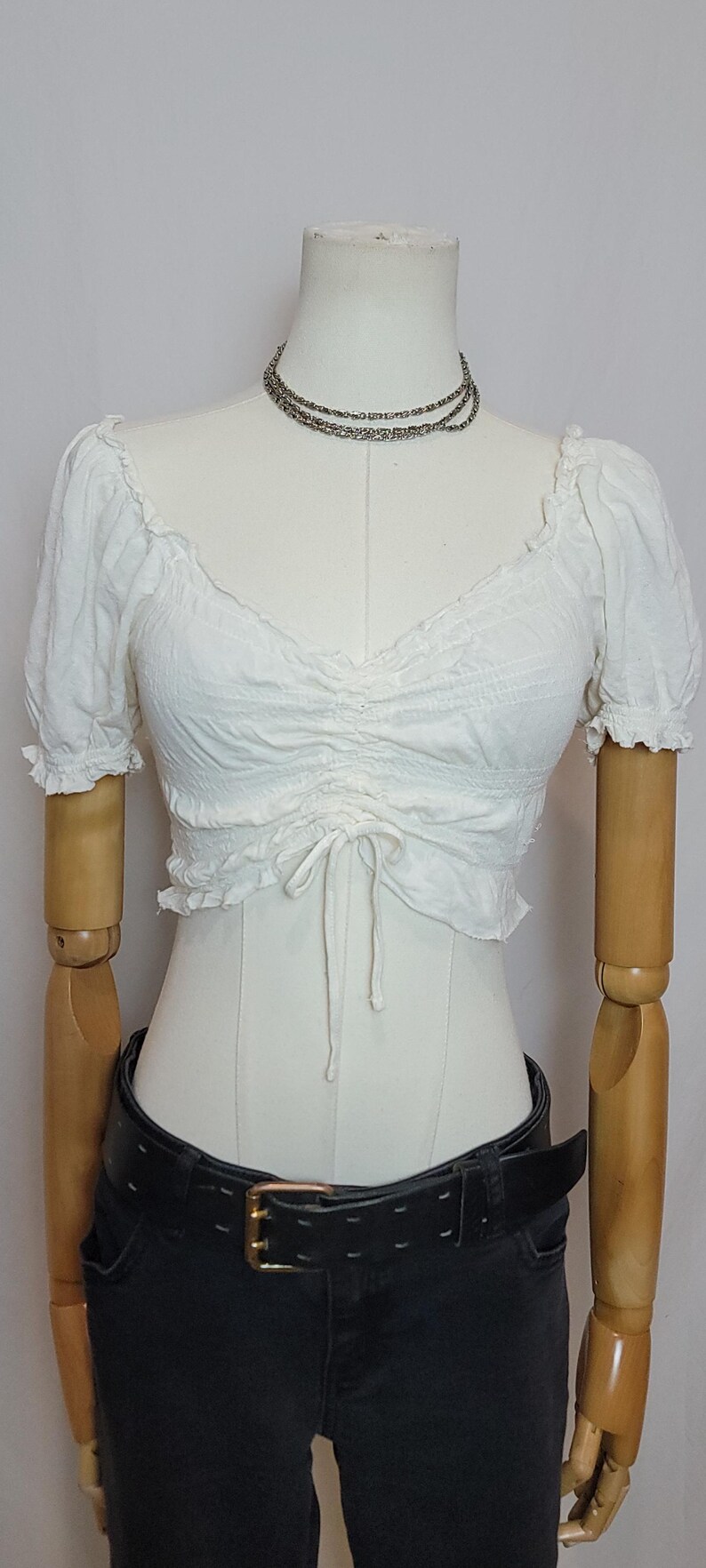 Op de afbeelding: Witte, gerimpelde crop top met korte mouwen en een trekkoord. Gedragen met een zwarte jeans en een zwarte riem met een gouden gesp. Een zilveren ketting is ook zichtbaar. De outfit wordt op een paspop getoond.