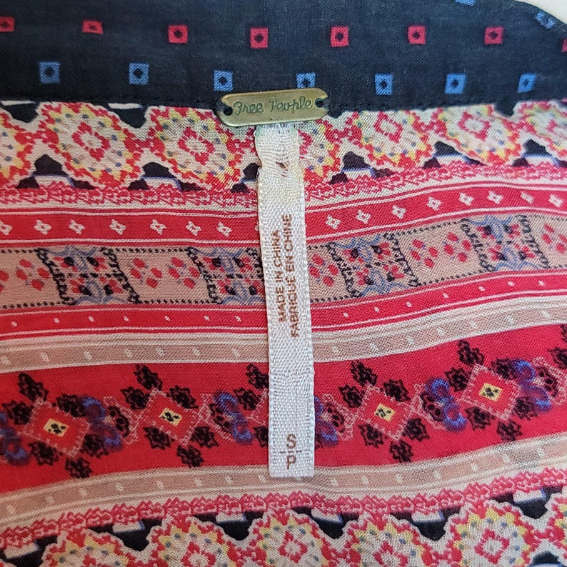 May include: Close-up of a patterned top with a navy blue collar featuring small red and blue squares. The top has a red, beige, and blue geometric design. A tag reads "Free People" and "Made in China".
