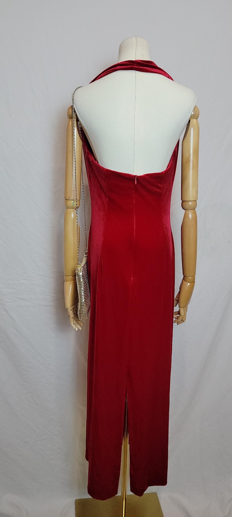 May include: A red velvet gown with a halter neckline and a back zipper. The dress has a floor-length skirt with a slit. A silver mesh purse hangs from the mannequin's arm.
