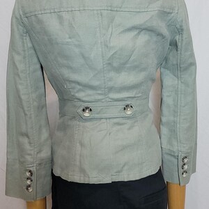 May include: A pale green blazer with three-quarter sleeves, a notched collar, and a belted waist. The blazer has silver buttons on the sleeves and waist. The jacket is displayed on a mannequin.