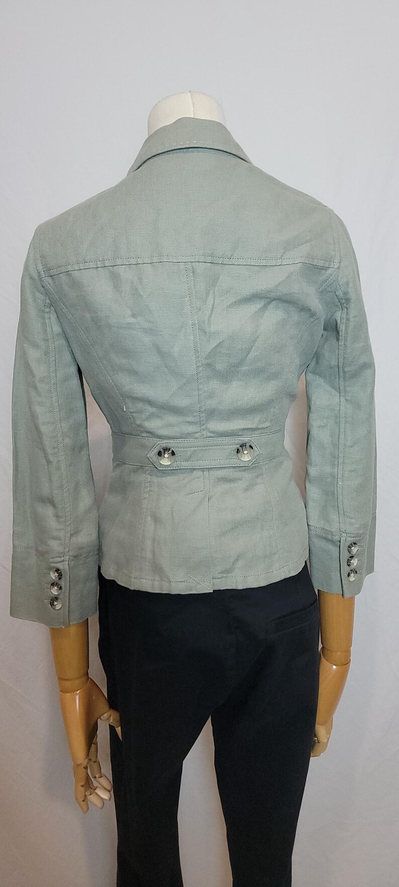 May include: A pale green blazer with three-quarter sleeves, a notched collar, and a belted waist. The blazer has silver buttons on the sleeves and waist. The jacket is displayed on a mannequin.