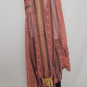 Puede incluir: Una blusa transparente de manga larga en coral y marr&oacute;n, con rayas verticales y detalles estampados. El escote tiene detalles bordados en azul y blanco. La blusa se usa con pantalones negros.