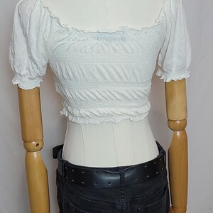 Op de afbeelding: Een witte crop top met korte, pofmouwen en een vierkante halslijn. De top is elastisch en wordt gedragen met een zwarte jeans en een zwarte riem. Een zilveren ketting is zichtbaar.