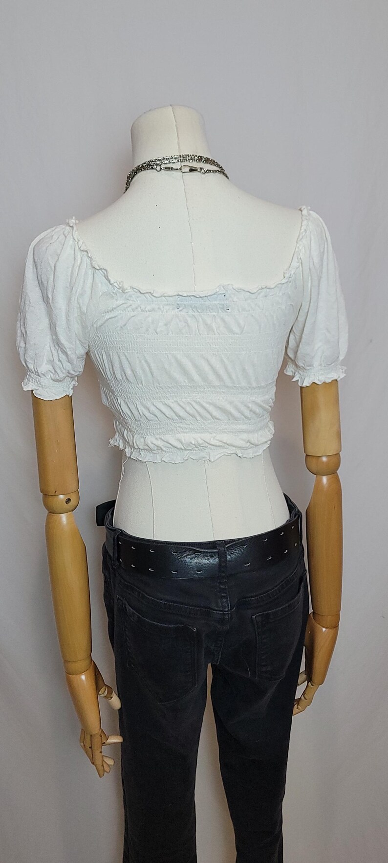 Op de afbeelding: Een witte crop top met korte, pofmouwen en een vierkante halslijn. De top is elastisch en wordt gedragen met een zwarte jeans en een zwarte riem. Een zilveren ketting is zichtbaar.