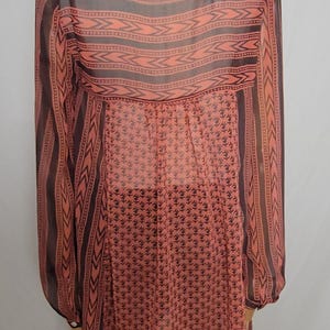 Puede incluir: T&uacute;nica transparente de color coral con mangas largas, exhibida en un maniqu&iacute;. La parte superior presenta un patr&oacute;n geom&eacute;trico en marr&oacute;n oscuro en los hombros y las mangas, con un patr&oacute;n floral repetido en la mitad inferior.