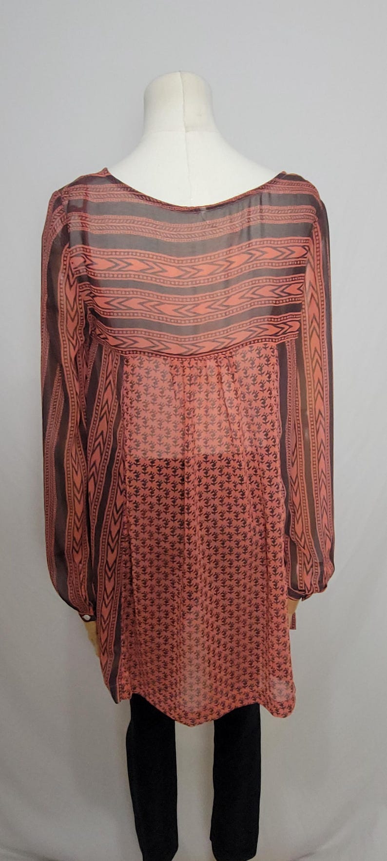 Puede incluir: T&uacute;nica transparente de color coral con mangas largas, exhibida en un maniqu&iacute;. La parte superior presenta un patr&oacute;n geom&eacute;trico en marr&oacute;n oscuro en los hombros y las mangas, con un patr&oacute;n floral repetido en la mitad inferior.