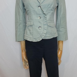 May include: A pale green blazer with three buttons, two front pockets, and a frilled detail. The blazer is worn with black trousers. The outfit is displayed on a mannequin.
