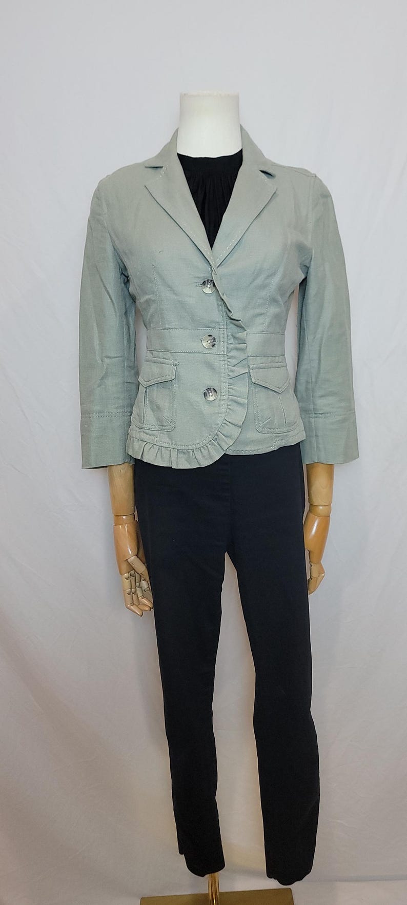 May include: A pale green blazer with three buttons, two front pockets, and a frilled detail. The blazer is worn with black trousers. The outfit is displayed on a mannequin.