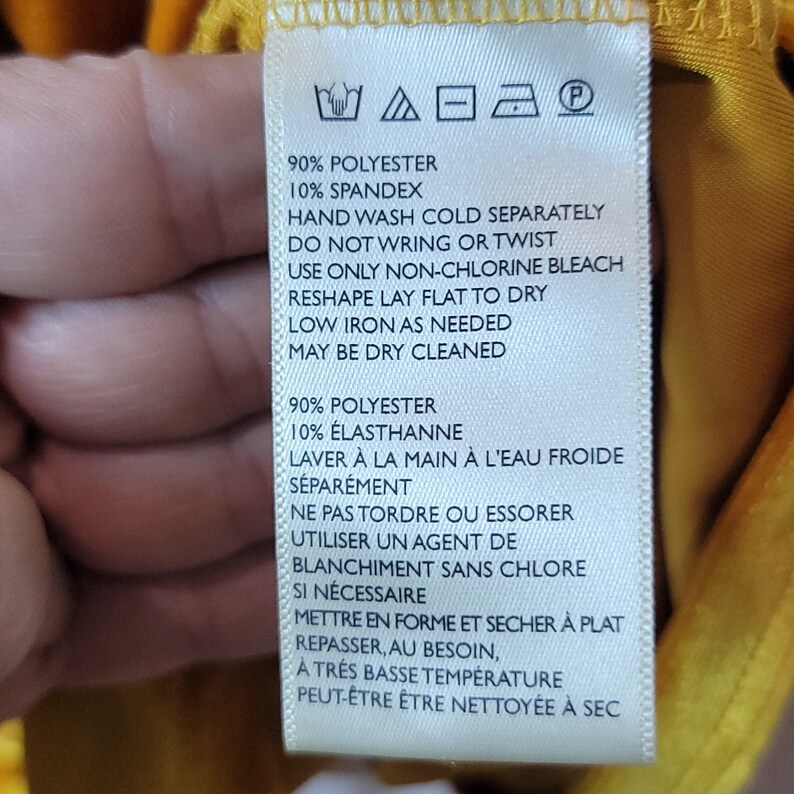 May include: A white clothing tag with black text, featuring washing instructions. The tag lists fabric composition as 90% polyester and 10% spandex. Instructions include hand washing in cold water, no wringing, and low-temperature ironing.