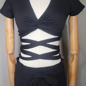 May include: A black crop top with a knotted front and criss-cross straps at the waist. The top is paired with black high-waisted trousers.