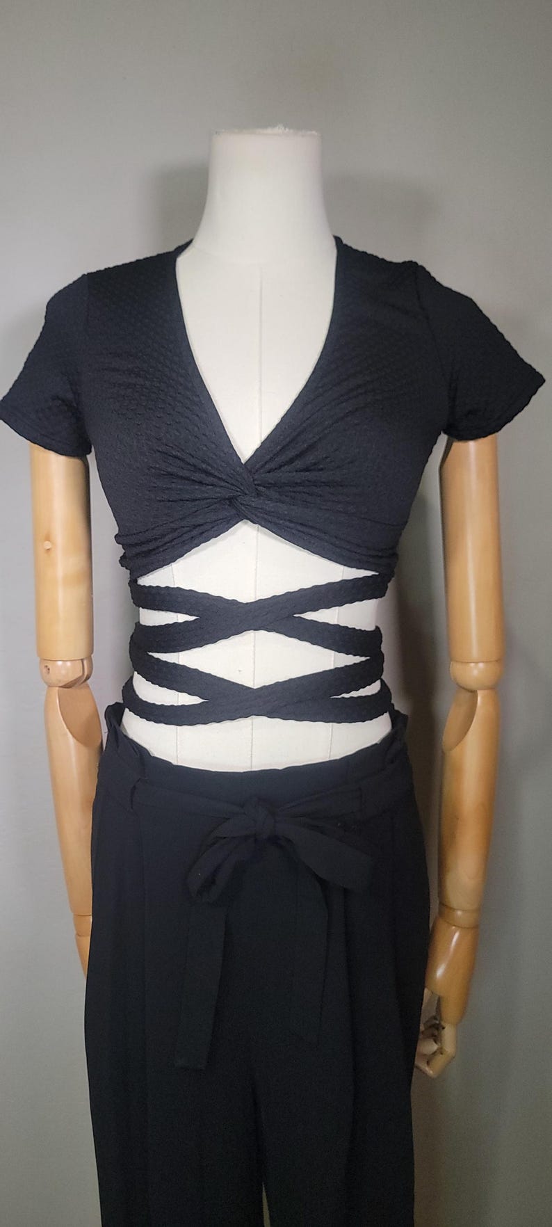 May include: A black crop top with a knotted front and criss-cross straps at the waist. The top is paired with black high-waisted trousers.