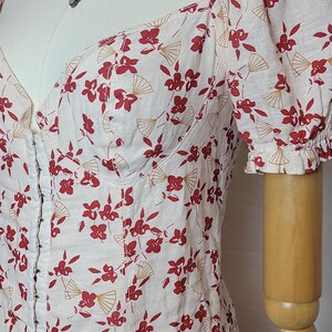 Puede incluir: Vestido rosa claro con estampado floral y abanicos rojos. El vestido tiene un escote coraz&oacute;n, mangas cortas abullonadas y cierre de corchetes. Se ve un collar plateado con un peque&ntilde;o colgante.