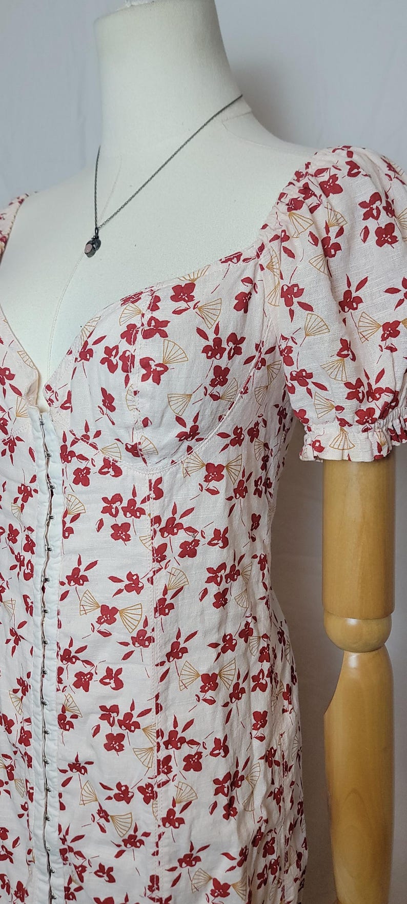 Puede incluir: Vestido rosa claro con estampado floral y abanicos rojos. El vestido tiene un escote coraz&oacute;n, mangas cortas abullonadas y cierre de corchetes. Se ve un collar plateado con un peque&ntilde;o colgante.