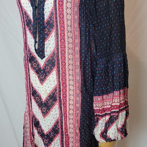 May include: A long-sleeved tunic top with a V-neckline. The top features a mix of patterns in navy, white, and pink. The sleeves are long and flared, with patterned cuffs. The fabric appears lightweight and flowing, suitable for warm weather.