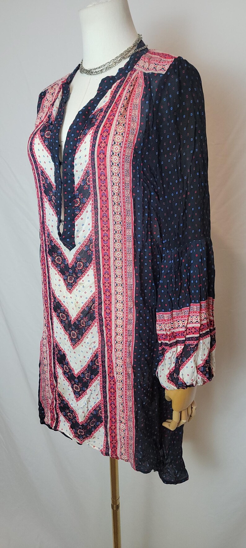May include: A long-sleeved tunic top with a V-neckline. The top features a mix of patterns in navy, white, and pink. The sleeves are long and flared, with patterned cuffs. The fabric appears lightweight and flowing, suitable for warm weather.