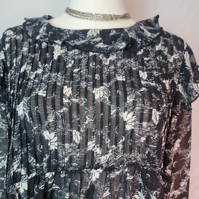 Puede incluir: Una blusa azul marino oscuro con un estampado floral blanco. La blusa presenta un corpi&ntilde;o plisado, un cuello con volantes y mangas largas. Un collar de gargantilla plateado es visible en el escote. La tela parece ser semitransparente.