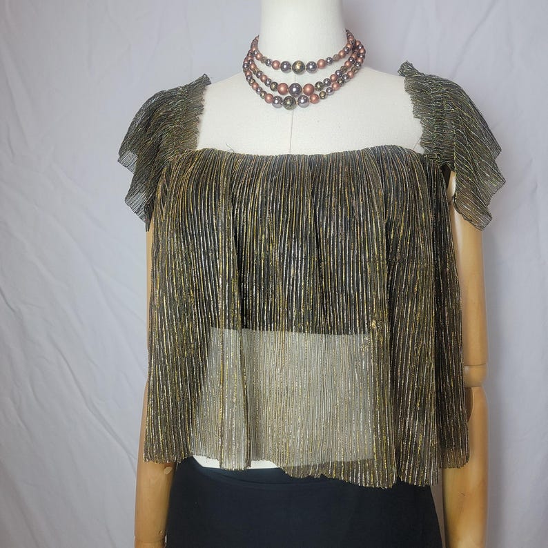 May include: A gold and black pleated top with short sleeves is displayed on a mannequin. The top has a square neckline and a layered design. A multi-strand beaded necklace in shades of brown and bronze is also visible.