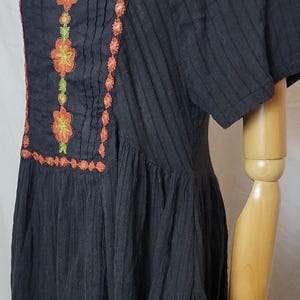 Puede incluir: Vestido negro con bordados florales naranjas y verdes. El vestido presenta un escote cuadrado y mangas cortas. El bordado est&aacute; alrededor del escote, en la parte delantera y a lo largo del dobladillo. El vestido se exhibe en un maniqu&iacute;.