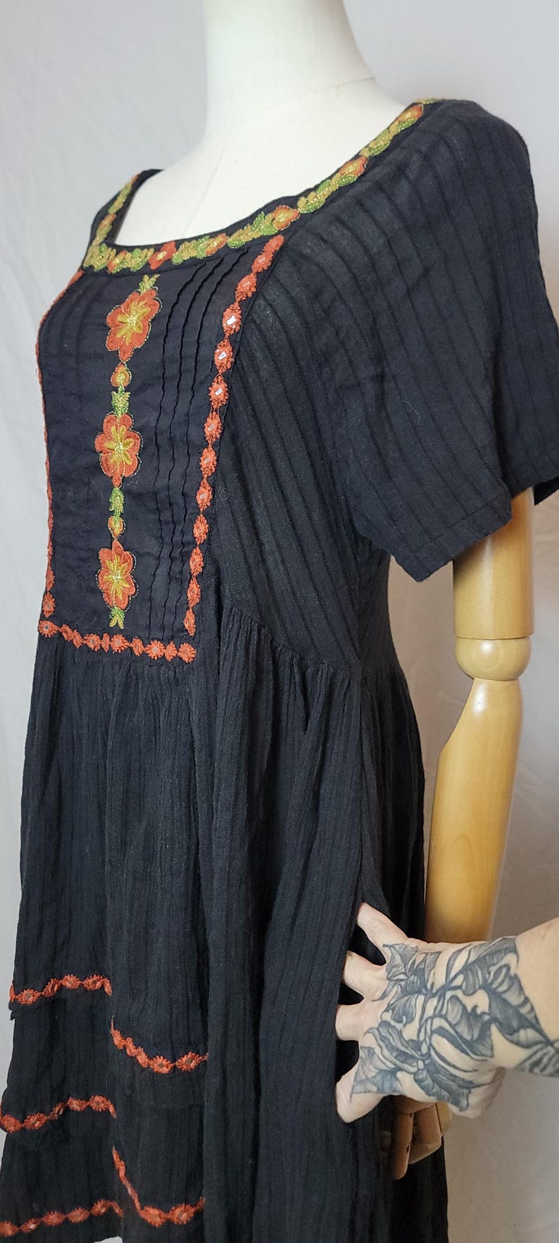 Puede incluir: Vestido negro con bordados florales naranjas y verdes. El vestido presenta un escote cuadrado y mangas cortas. El bordado est&aacute; alrededor del escote, en la parte delantera y a lo largo del dobladillo. El vestido se exhibe en un maniqu&iacute;.