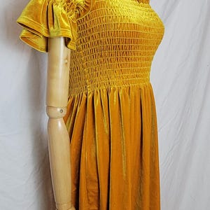 May include: Mustard yellow velvet dress with a square neckline and ruffled sleeves. The bodice features a smocked design, and the dress has a flared skirt. A floral necklace is visible. The dress is displayed on a mannequin.