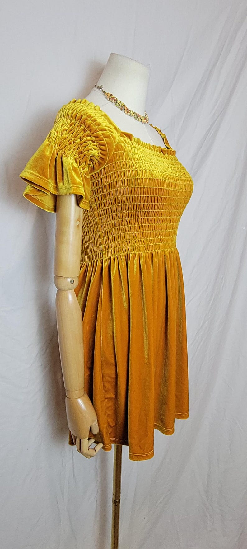 May include: Mustard yellow velvet dress with a square neckline and ruffled sleeves. The bodice features a smocked design, and the dress has a flared skirt. A floral necklace is visible. The dress is displayed on a mannequin.