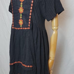 Puede incluir: Vestido negro con mangas cortas y bordado floral. El vestido presenta bordados florales en naranja y verde alrededor del escote y en la parte delantera. El vestido se exhibe en un maniqu&iacute;.