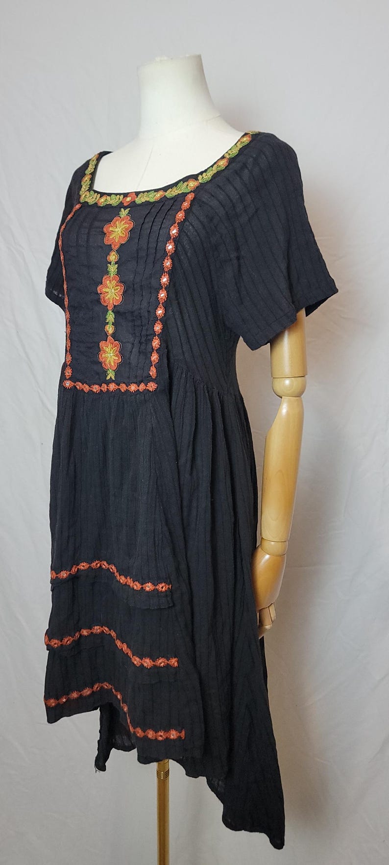 Puede incluir: Vestido negro con mangas cortas y bordado floral. El vestido presenta bordados florales en naranja y verde alrededor del escote y en la parte delantera. El vestido se exhibe en un maniqu&iacute;.