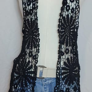 May include: A sleeveless black crochet vest featuring a repeating floral pattern with openwork detailing. The vest is worn over a white belt and light blue denim shorts, with a silver-toned chain necklace visible at the neckline.