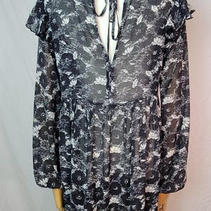 Puede incluir: Vestido con estampado floral en blanco y negro con mangas largas y cuello con volantes. El vestido tiene una silueta fluida y una espalda con abertura de cerradura con cierre de lazo. La tela parece ligera y semitransparente.