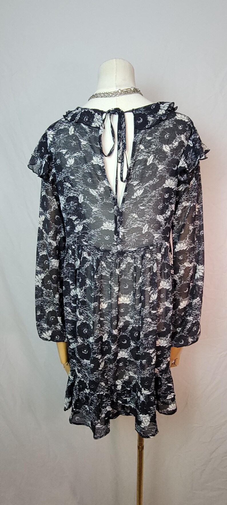 Puede incluir: Vestido con estampado floral en blanco y negro con mangas largas y cuello con volantes. El vestido tiene una silueta fluida y una espalda con abertura de cerradura con cierre de lazo. La tela parece ligera y semitransparente.