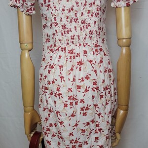 Puede incluir: Vestido blanco con estampado floral rojo y escote cuadrado. El vestido tiene mangas cortas y un corpi&ntilde;o ajustado. Un bolso de mano rojo es visible en la imagen.