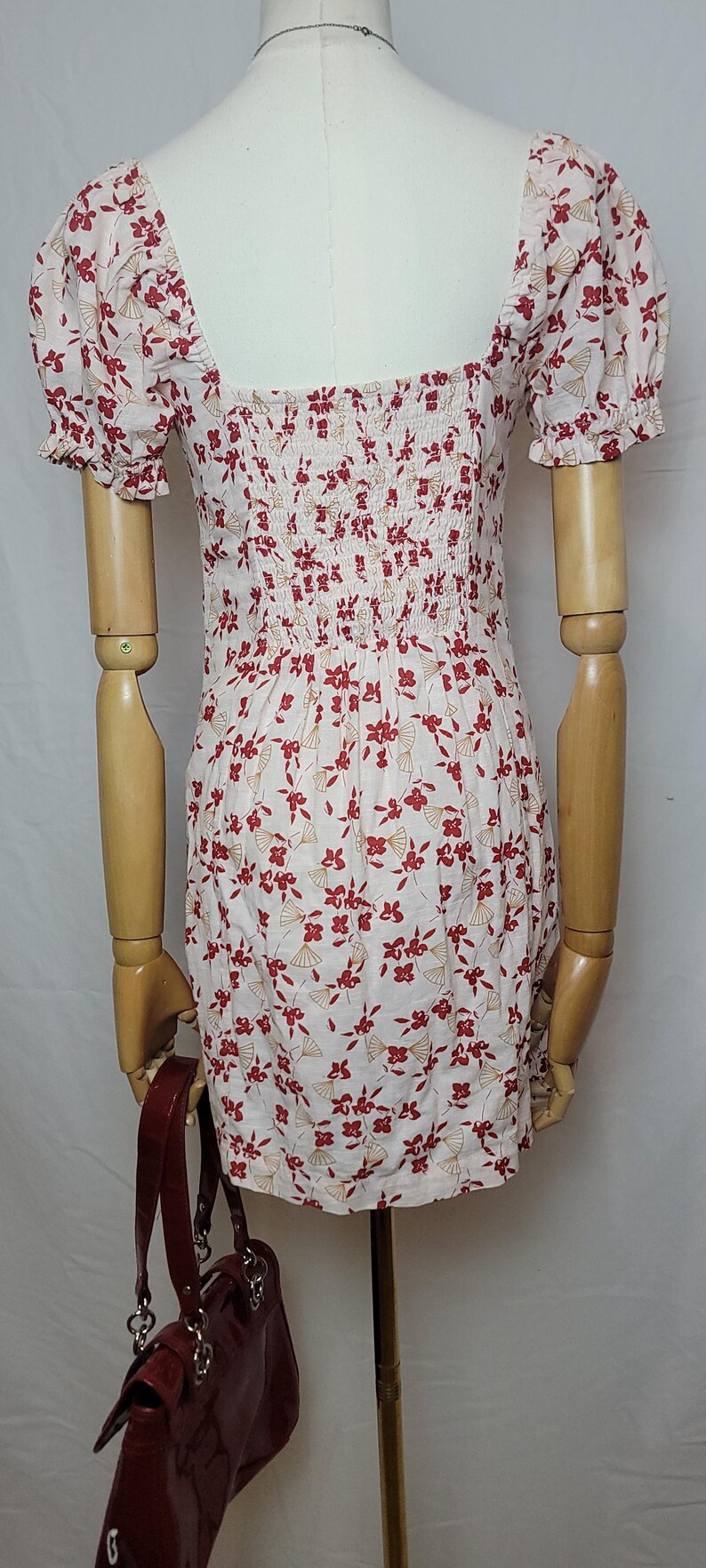 Puede incluir: Vestido blanco con estampado floral rojo y escote cuadrado. El vestido tiene mangas cortas y un corpi&ntilde;o ajustado. Un bolso de mano rojo es visible en la imagen.